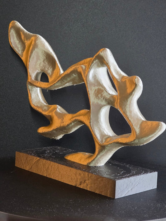 An abstract sculptural wall art piece with flowing organic shapes in metallic gold and silver, mounted on a rectangular dark base against a dark gray wall.