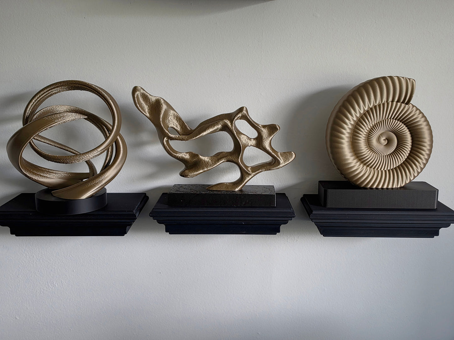 Three decorative wall sculptures displayed on black pedestals against a plain light gray wall: an abstract interlocking ring design, a fluid organic branch-like form, and a spiral ammonite-style shell sculpture.