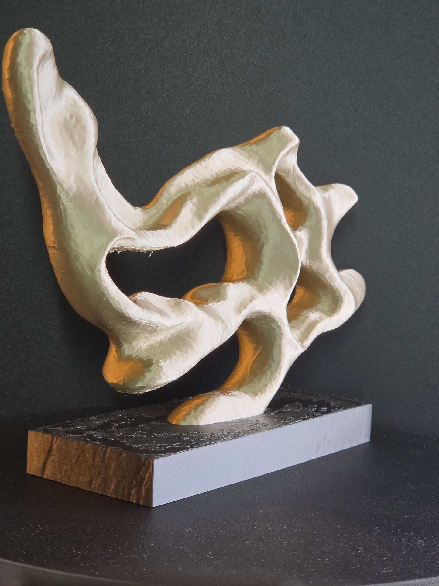Abstract sculptural object with flowing organic folds in off-white and pale green tones, displayed on a rectangular base against a dark charcoal background.