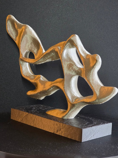 An abstract sculptural wall art piece with flowing organic shapes in metallic gold and silver, mounted on a rectangular dark base against a dark gray wall.
