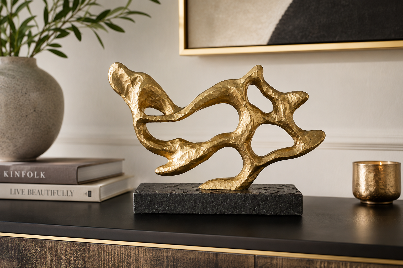Abstract gold metal sculpture on a black rectangular base, styled on a dark console table with a vase, books, and a small metallic cup in a home decor setting.