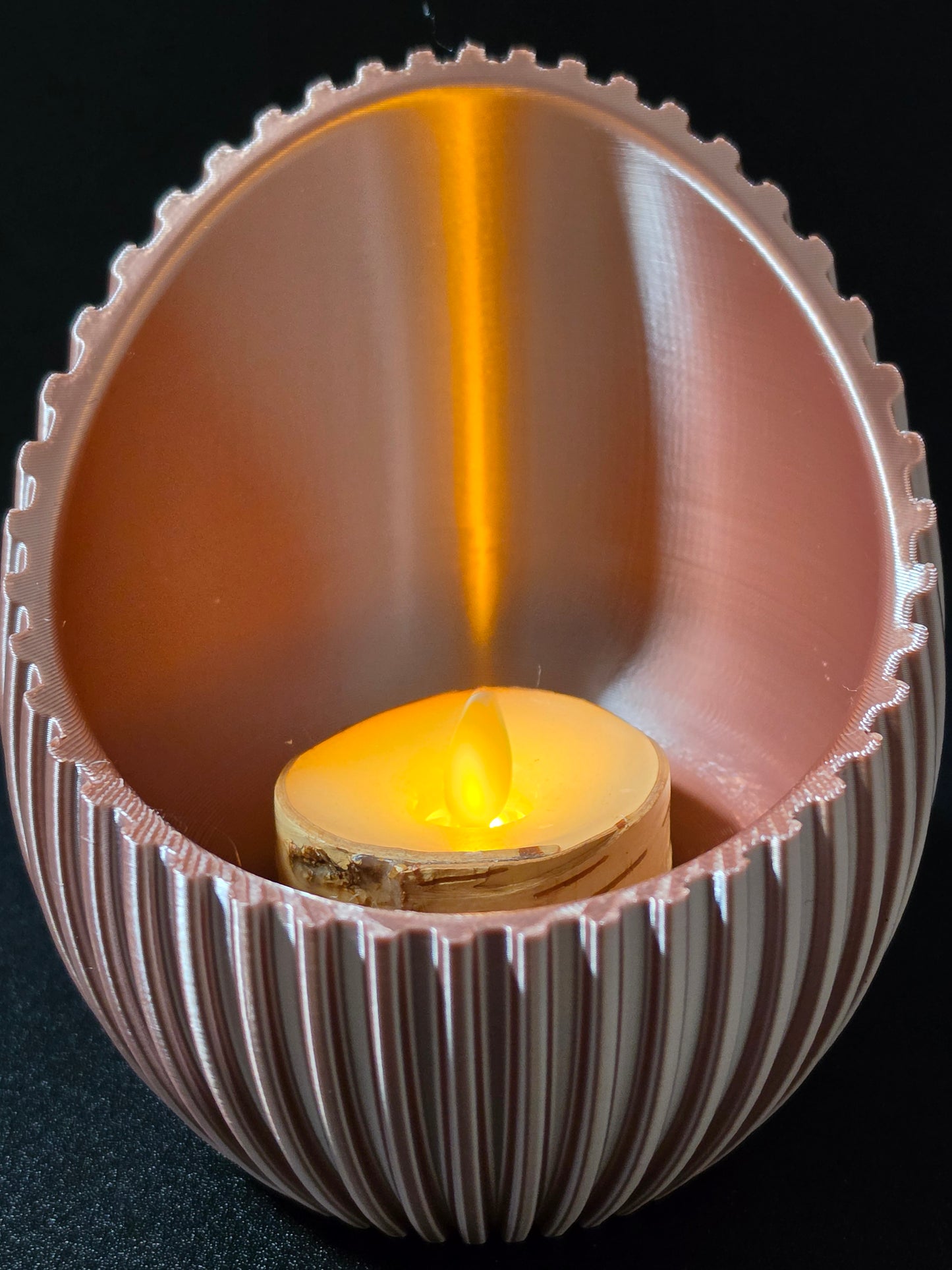 A close-up of a ribbed, egg-shaped candle holder in a warm metallic rose gold finish, containing a small lit tealight candle that casts a bright yellow glow against a dark background.