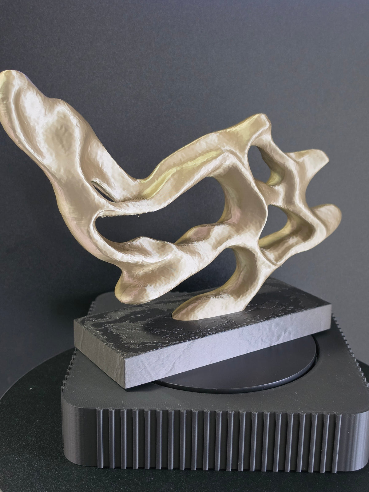 Abstract sculptural art piece in a metallic gold finish, black art display base against a dark gray background.