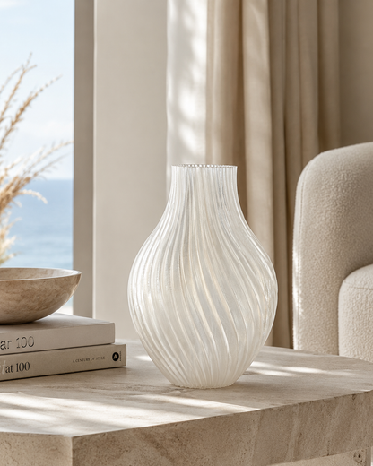 White textured vase on a table with books and a bowl in a room with curtains and a window.